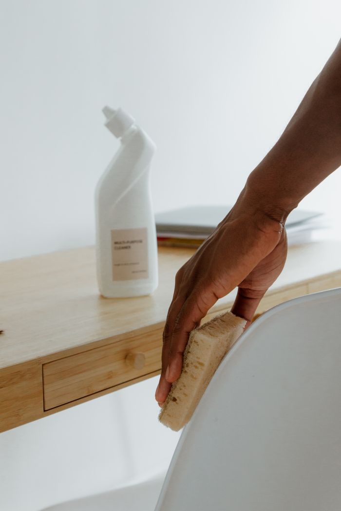 A pared-back room shows a person wiping down a chair with a sponge and a cleaning solution.
